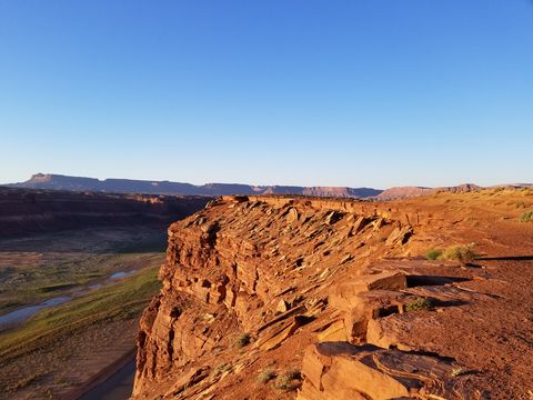 Early sunlight on a red sandstone rim dropping to a green river bottom, with flat-topped buttes along the horizon.