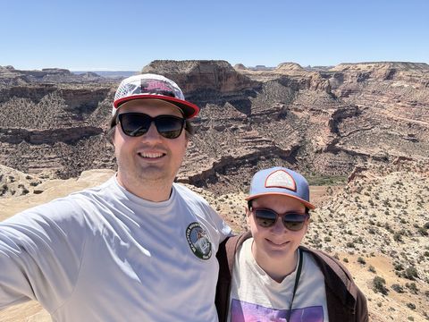 Two people in sunglasses and ball caps smiling for a selfie at the canyon rim, the layered canyon spread out behind them.