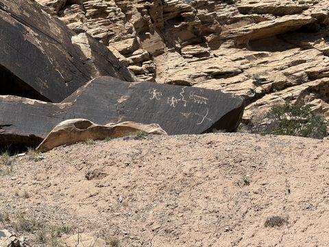 The same petroglyph boulder from farther back, its pecked figures small against the tilted slab and the broken cliff above.
