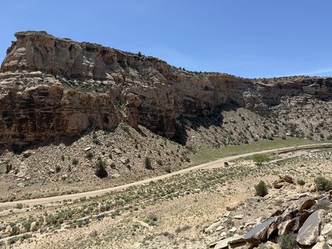 A long banded cliff above a talus slope, with the dirt road and a vehicle small along its base.