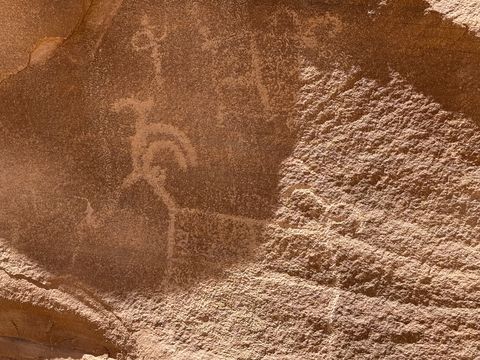 A close view of pecked petroglyphs on a varnished rock face, including a figure with a curved headdress and a horned animal.