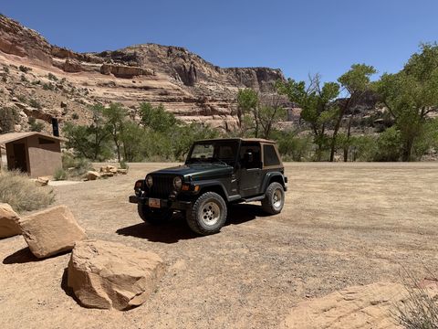 The Jeep parked on a gravel flat below a tall banded cliff, cottonwoods and a vault toilet in the background.