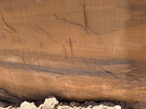 A tan sandstone wall with a faint dark red painted figure near its center, above a horizontal band of desert varnish.