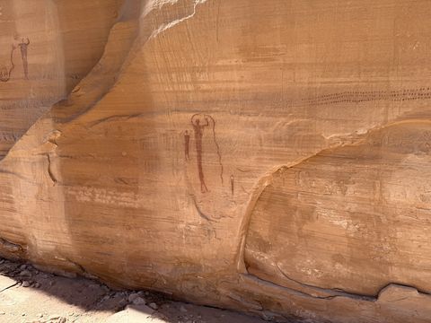 Slender dark red painted human-like figures on a smooth sandstone wall, one with raised arms.