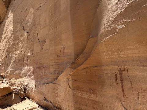 A sunlit stretch of sandstone wall with several red painted figures spaced along it, one pair with arms raised.