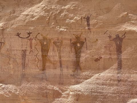 A pictograph panel of tall, broad-shouldered human-like figures painted in red and brown, some with horned headdresses and patterned bodies.