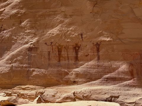 The pictograph panel seen from farther back, its row of standing figures small against the pale sandstone alcove wall.