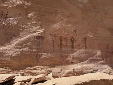 Four tall painted figures in a row on the cliff face, weathered and faded, above a bench of broken sandstone.