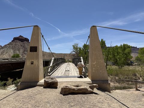 A wooden-decked suspension footbridge between two concrete towers, a person standing partway across, red cliffs on the far bank.