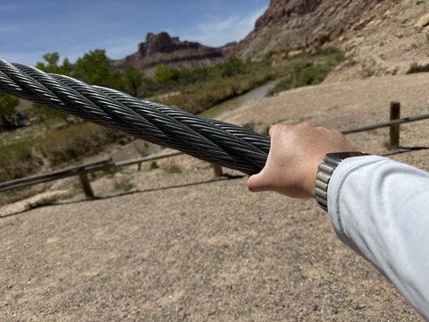 A hand gripping one of the bridge's thick steel suspension cables, the river and canyon walls out of focus behind.
