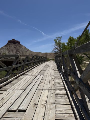 Looking down the plank deck of the swinging bridge between its timber railings, a dark-capped butte beyond the far end.