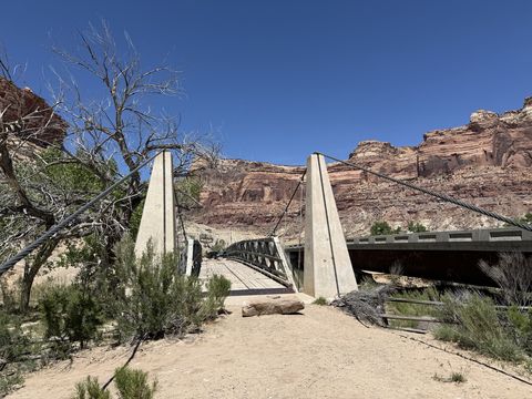 The suspension bridge seen from the bank, its two concrete towers and cables framed against the red canyon wall, with the modern road bridge behind it.