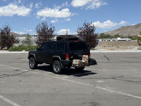 A black Jeep Cherokee loaded with a roof cargo box and a rear cargo carrier, parked alone in an empty asphalt lot with dry brown hills behind it.