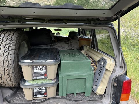 The open rear hatch of the Jeep showing camp gear packed to the roof: stacked tan storage totes, a green military-style trunk, a folding table on edge, bedding and a spare tire.