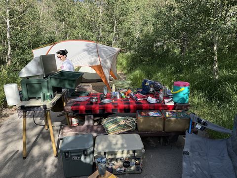 A set-up campsite: a dome tent under aspens, a camp kitchen table with a red plaid cloth covered in cookware and gear, a portable stove stand, and two hard coolers on the ground.