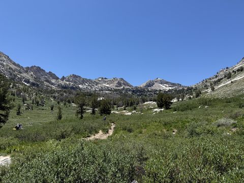 A dirt trail crossing a green alpine meadow toward a cirque of snow-streaked peaks, with hikers small on the path.