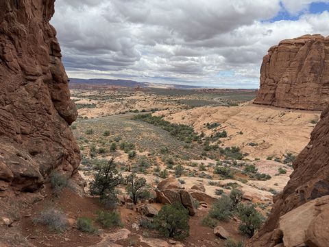A view out between two red sandstone walls to a wide slickrock basin dotted with juniper, storm clouds stacked above the far mesas.