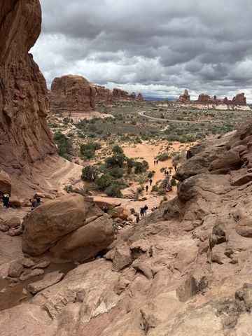 Looking out from inside a sandstone alcove over the Windows Section, visitors small on the sand trail below and dark clouds over the fins beyond.