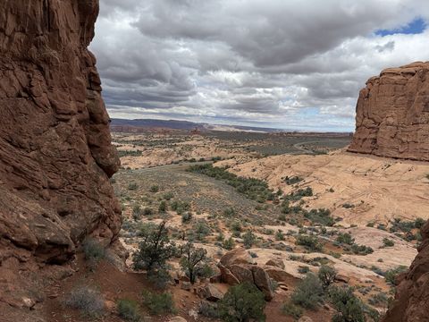 The same gap between two rock walls, framing a valley of slickrock and sagebrush with a distant road and cloud-shadowed cliffs.