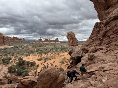 People scrambling on the boulders below a sandstone alcove, with sandstone fins and towers spread across the valley under a grey sky.