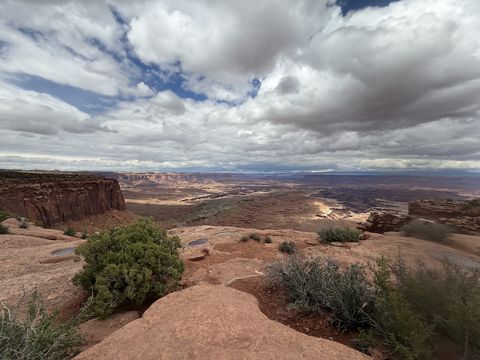A rim of red slickrock and juniper above a broad canyon, with a towering bank of grey and white cloud filling the sky.