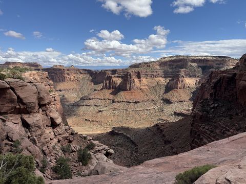 Shafer Canyon from the rim, its stepped red walls and talus slopes dropping to a canyon floor, white clouds scattered across blue sky.