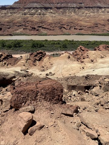 Broken red and white rock in the foreground, the Green River and its green banks below a long banded cliff wall.
