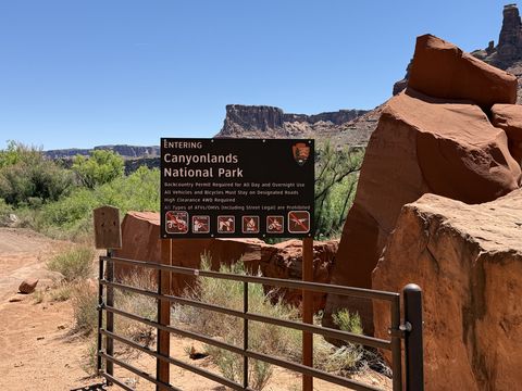 A brown park sign reading "Entering Canyonlands National Park" beside a closed metal gate on the dirt road, red cliffs behind.