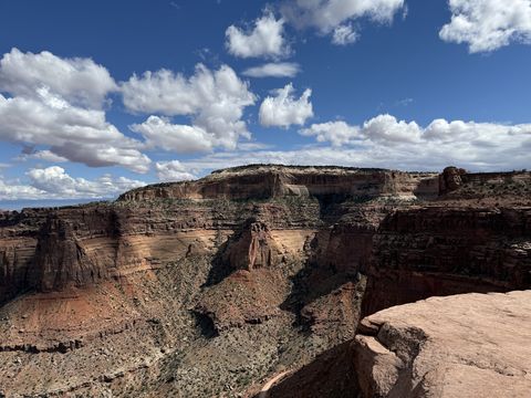 The banded cliffs across Shafer Canyon, layers of red and grey rock stepping up to a flat mesa top under cumulus clouds.