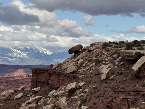 A slab of caprock balanced on the rim above a rubble slope, the snow-covered La Sal Mountains on the horizon behind.