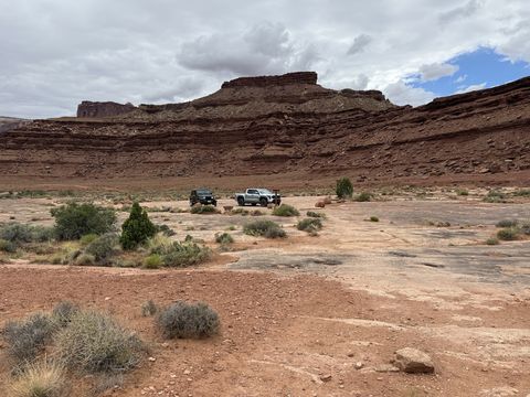 A Jeep and a pickup parked on a slickrock flat below a long banded cliff, storm clouds overhead.