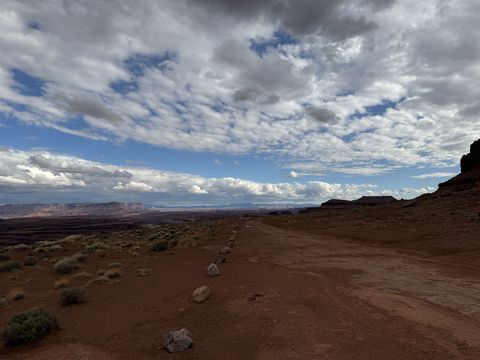 The White Rim road running away across open desert, cliffs on the right and a bright cloud-streaked sky above.