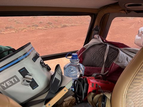 The loaded interior of the Jeep from the front seat: cooler, water bottle, stuffed packs and a red toolbox, desert beyond the glass.