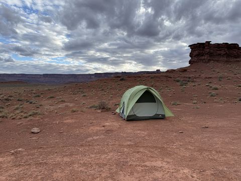 A green dome tent pitched alone on a red dirt flat, a small butte to the right and a heavy evening overcast above.
