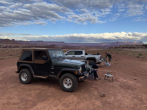 The Jeep and a pickup parked at camp, camp chairs and a folding table set out beside them, mesas on the horizon at dusk.