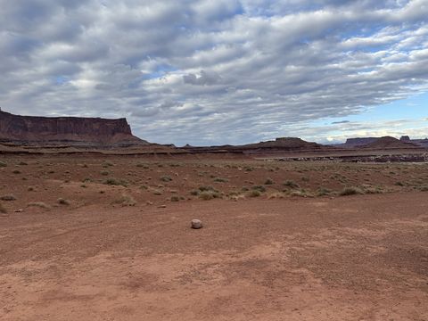A flat, empty campsite of red dirt and low brush, layered mesas standing along the skyline under a grey evening sky.
