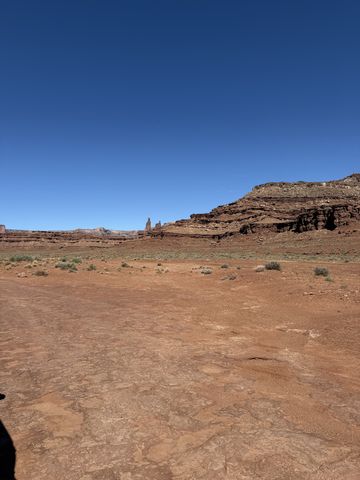 A flat pan of slickrock and red dirt, with Monster Tower and Washer Woman Arch small on the distant cliff line.