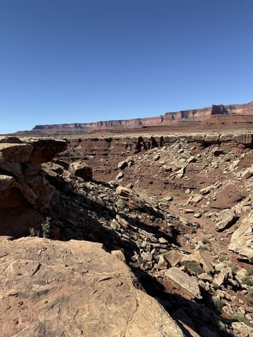A broken rim of red rock in the foreground, with a long flat-topped mesa across the canyon under a cloudless sky.