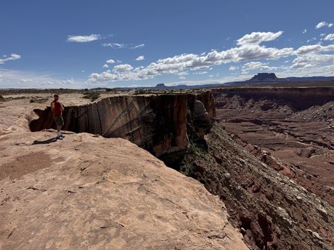 A person in a red shirt standing at the edge of a sheer-walled rim, a butte on the horizon behind them.