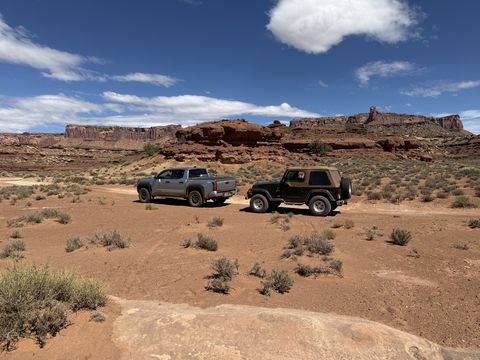 The Jeep and a pickup parked on a red dirt flat, low sandstone ledges and a broken skyline behind them.