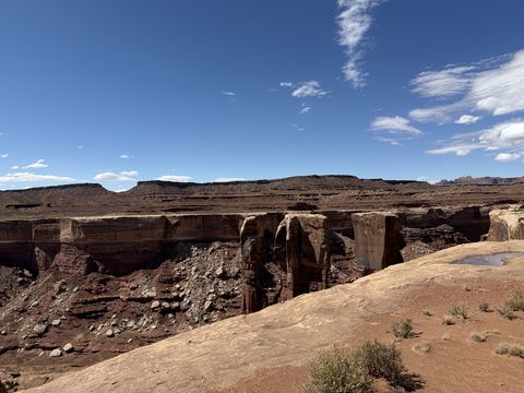A row of blocky red towers along the far wall of a side canyon, a pothole of water on the slickrock in front.
