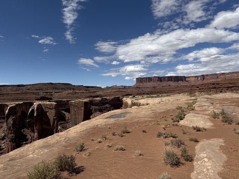 Slickrock and shallow potholes at the rim, with a long banded mesa across the canyon under scattered clouds.