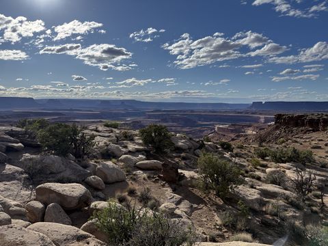 Boulders and juniper on a slickrock bench with the sun high above, canyon country receding to a flat horizon.