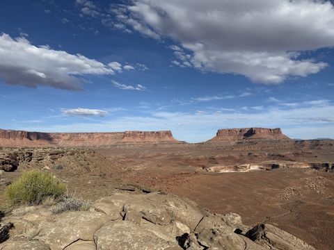 Two flat-topped mesas standing above a wide desert basin, seen across a rocky rim under scattered cloud.