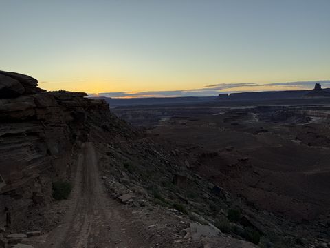 A narrow dirt road running along a steep ridge at sunset, the sky glowing orange over the dark canyon.