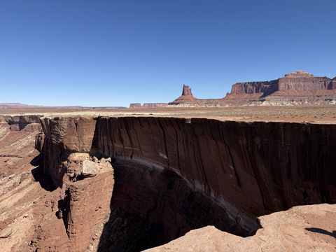 A sheer, dark red canyon wall curving below the rim, with a butte and a long mesa on the horizon.